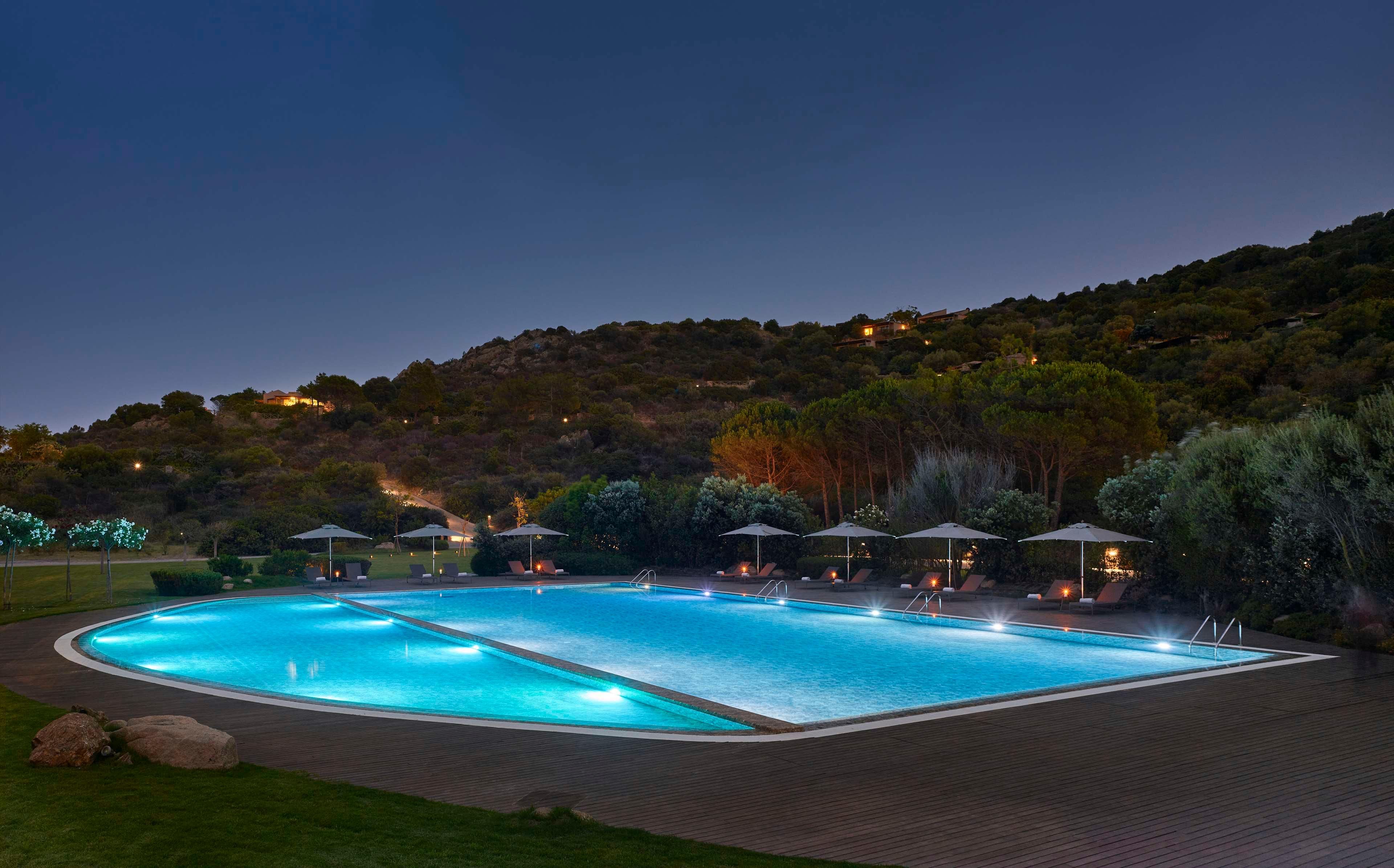 Una gran piscina al aire libre con numerosas tumbonas y sombrillas, rodeada por un jardín ajardinado y una colina de fondo.