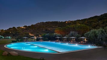 Una gran piscina al aire libre con numerosas tumbonas y sombrillas, rodeada por un jardín ajardinado y una colina de fondo.
