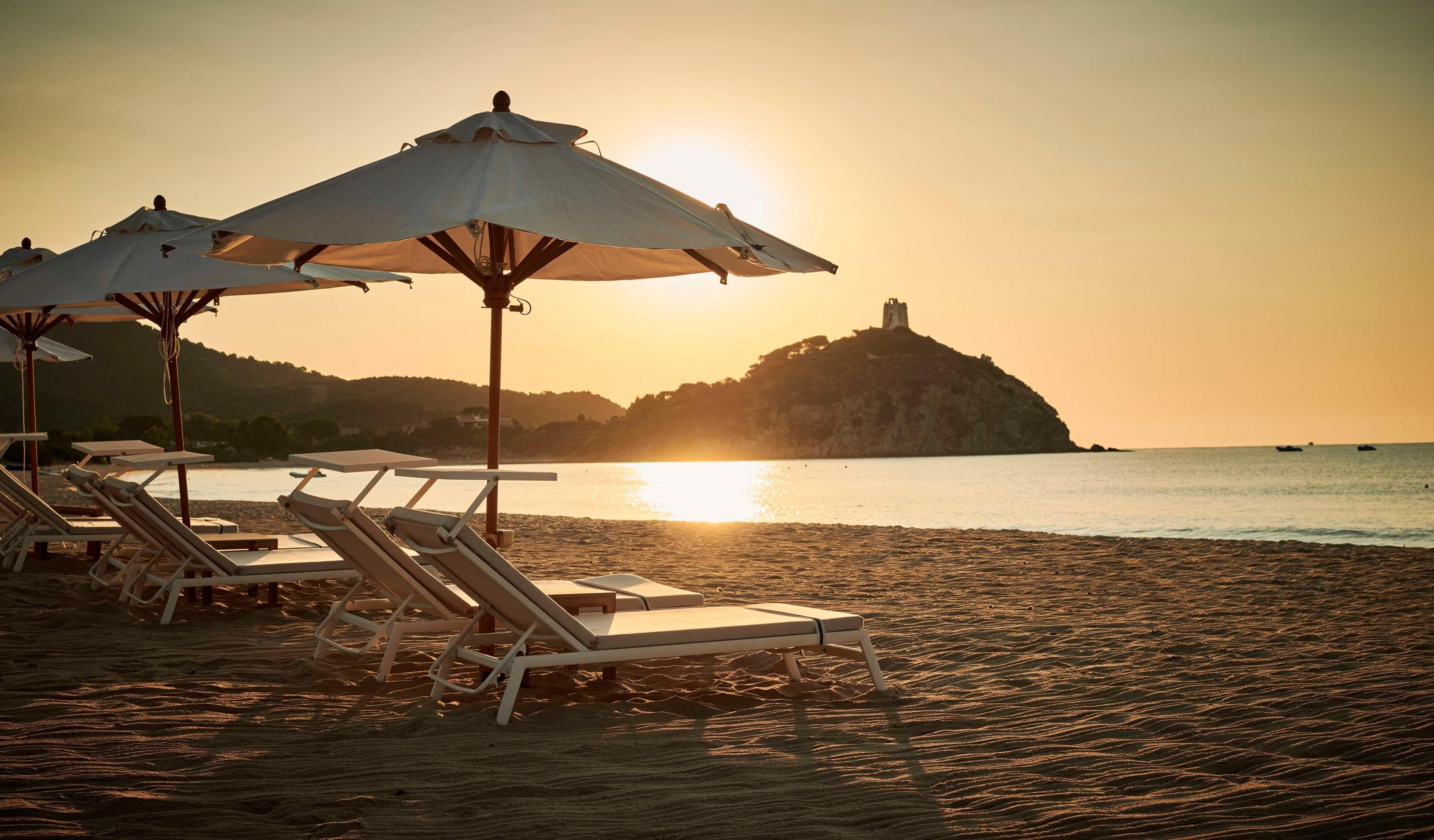 Une plage avec des transats et des parasols, une mer calme et, au loin, sur une colline, un château.