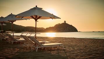 Une plage avec des transats et des parasols, une mer calme et, au loin, sur une colline, un château.