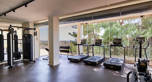 Un gimnasio con cintas de correr, mancuernas y máquinas de pesas, con ventanas amplias y vistas a árboles.