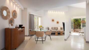 Un salon moderne avec une table à manger en verre, des chaises en osier, un canapé et une table basse. On y trouve trois miroirs fixés au mur et un meuble en bois.