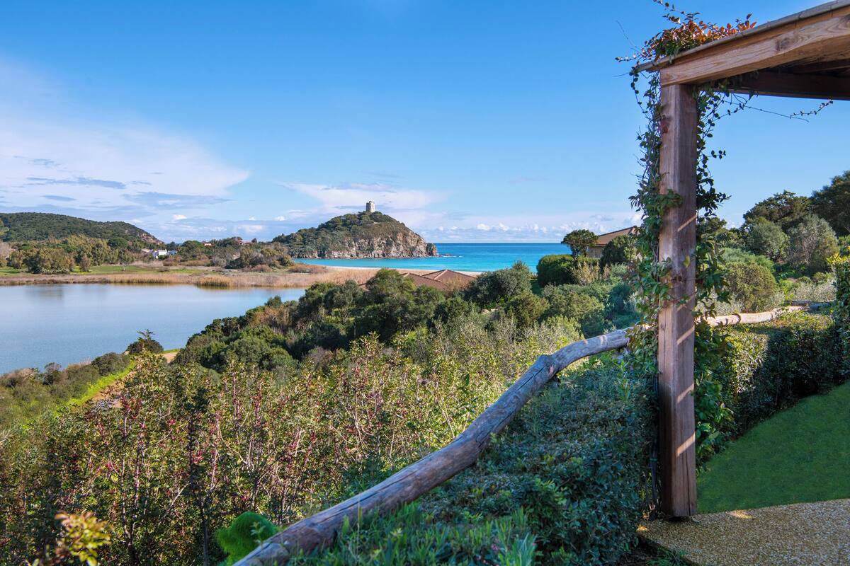 Un paysage côtier avec une pergola en bois, une végétation luxuriante et une vue lointaine d’une colline surmontée d’une construction.