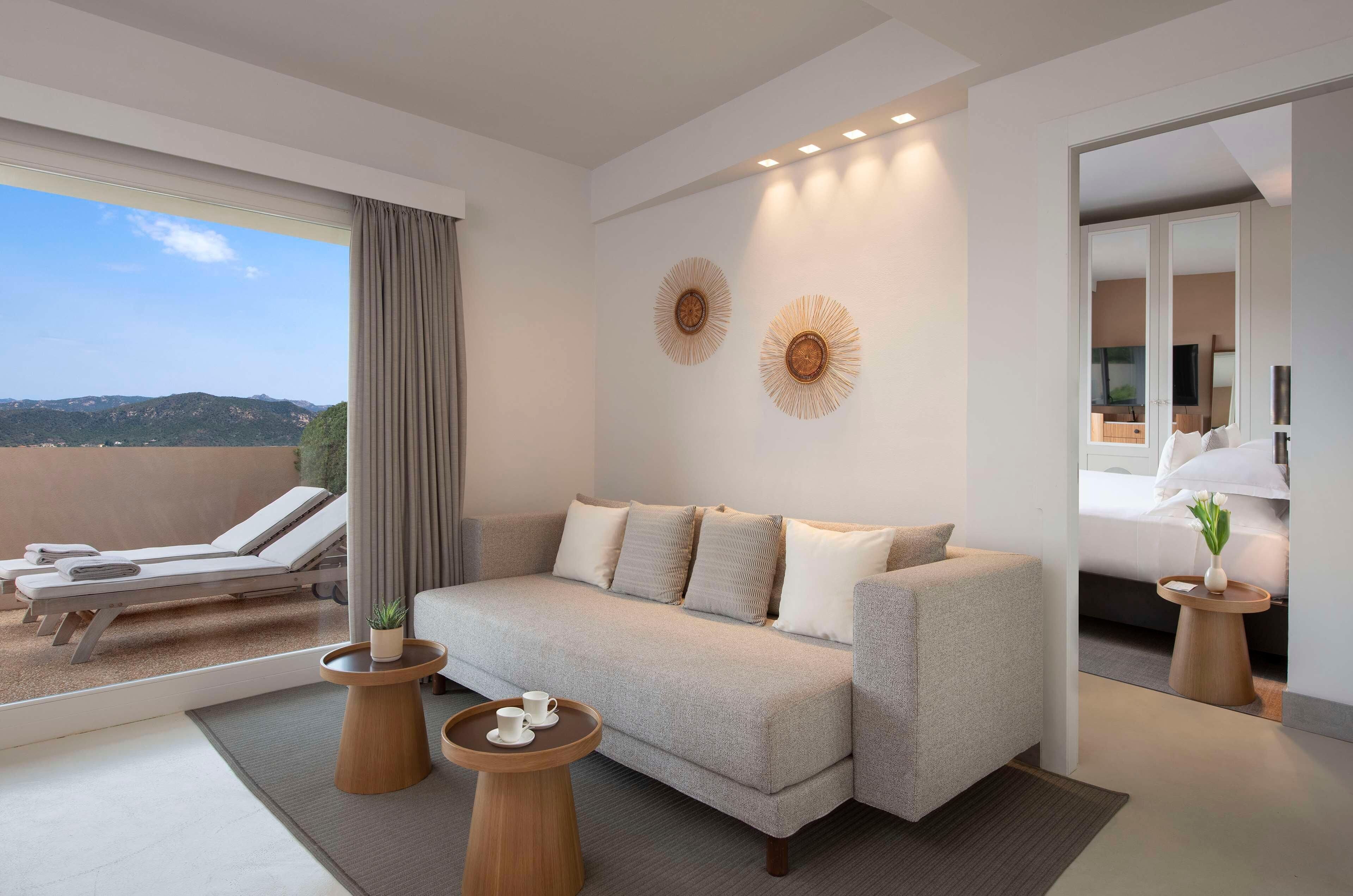 Une chambre d’hôtel moderne avec un canapé, deux tables d’appoint, une petite table ronde avec des tasses, une plante en pot et une vue sur l’extérieur.