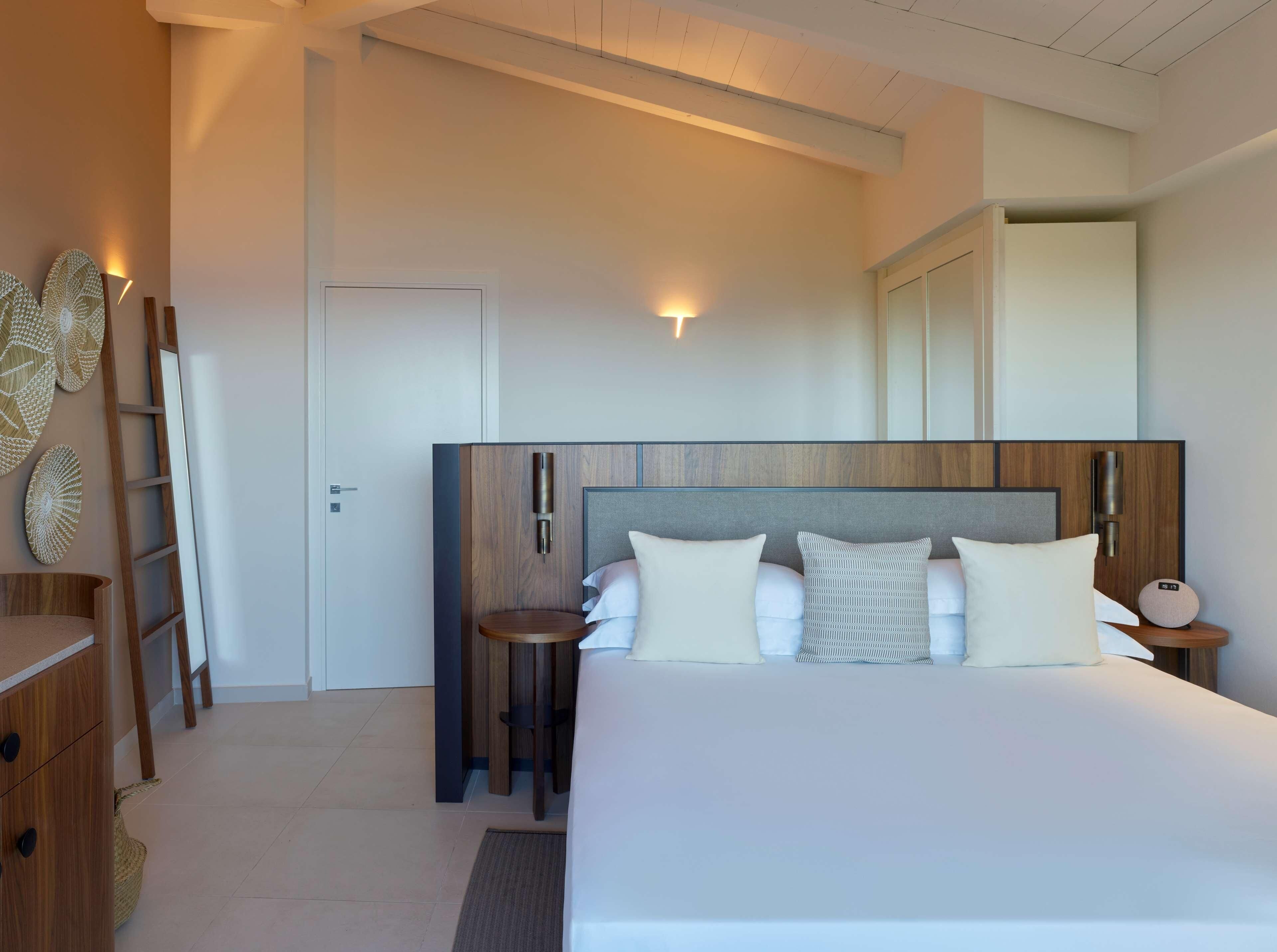 A modern hotel room with a large bed, bedside tables, a wooden headboard, and wall-mounted lighting.