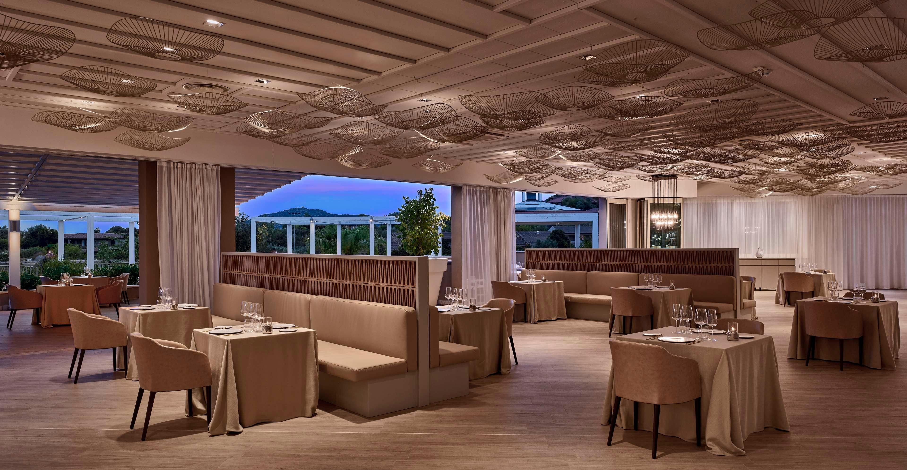 A modern dining area with booths and tables, large windows with curtains, and a ceiling with unique light fixtures.