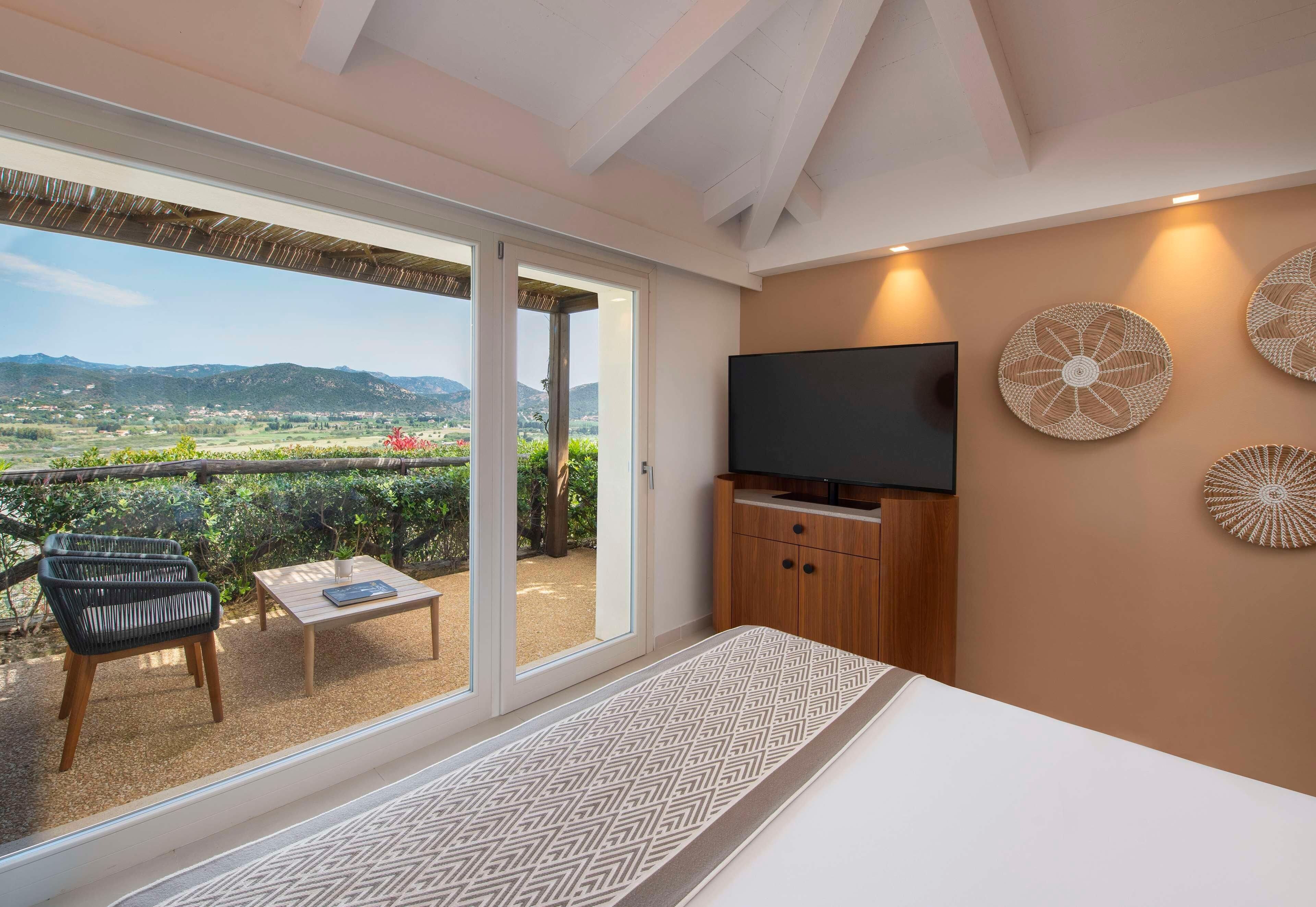 Una habitación con cama, televisor, mesa y silla. Hay una puerta corrediza de vidrio que da a un espacio exterior con vistas al campo.