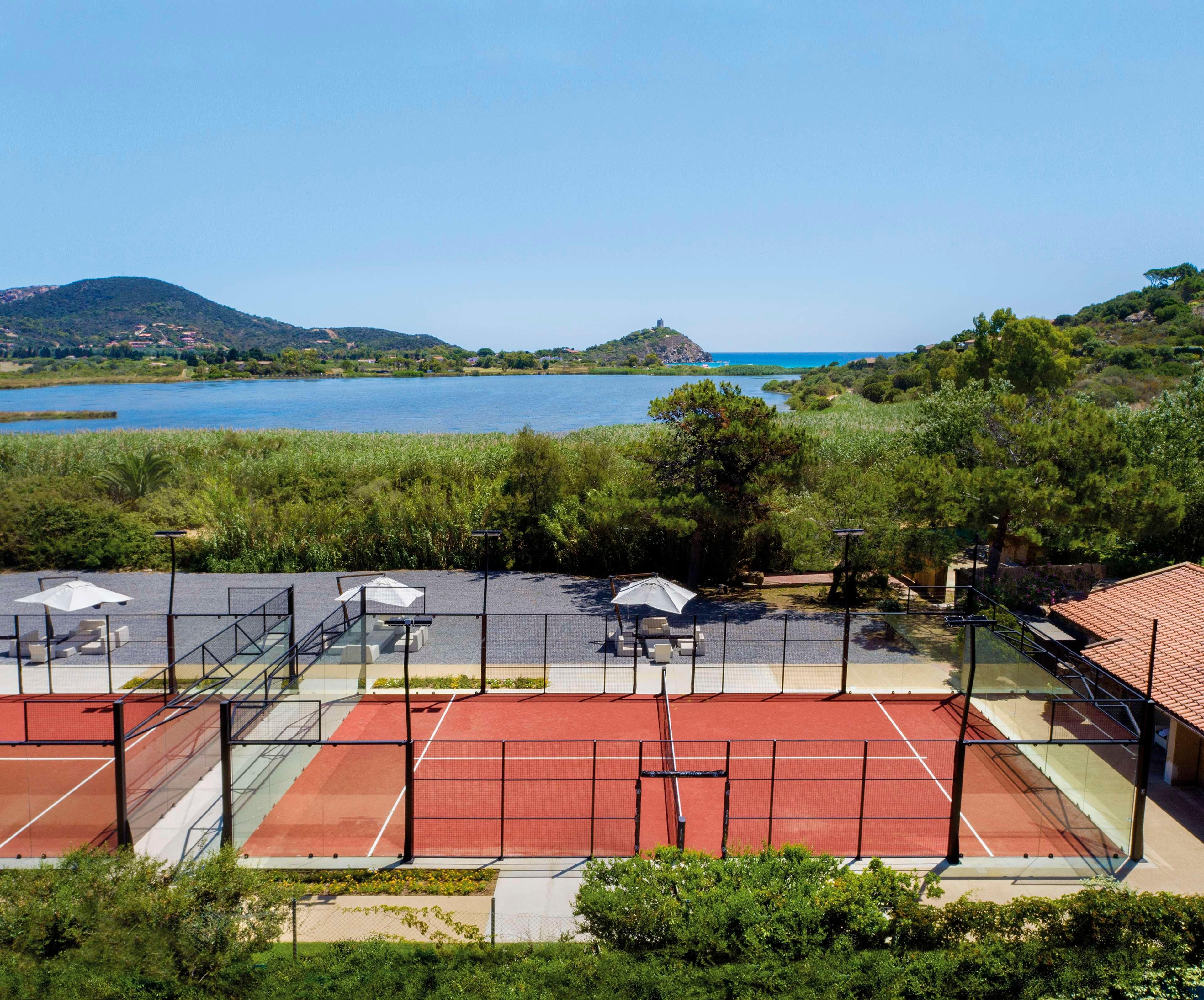 A tennis court with a net, surrounded by greenery and a view of a lake and hills.