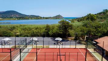 A tennis court with a net, surrounded by greenery and a view of a lake and hills.