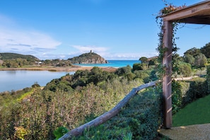 Paisaje costero con una pérgola de madera, abundante vegetación y una vista distante de una colina con una edificación en la cima.