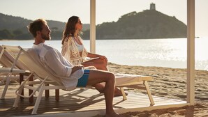 Un couple est assis sur des chaises longues en bois, sur une terrasse donnant sur une étendue d’eau, avec une colline en arrière-plan.