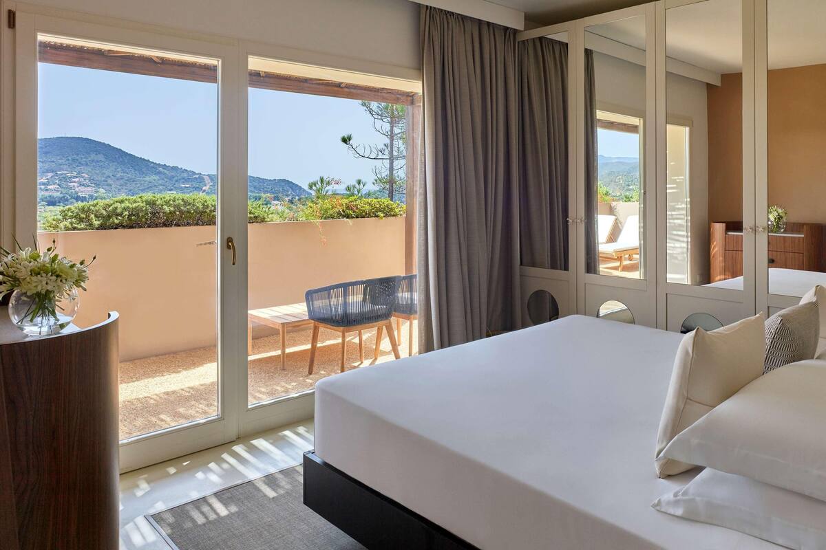 Une chambre d’hôtel avec un grand lit, un balcon donnant sur les collines et un bureau avec une chaise.