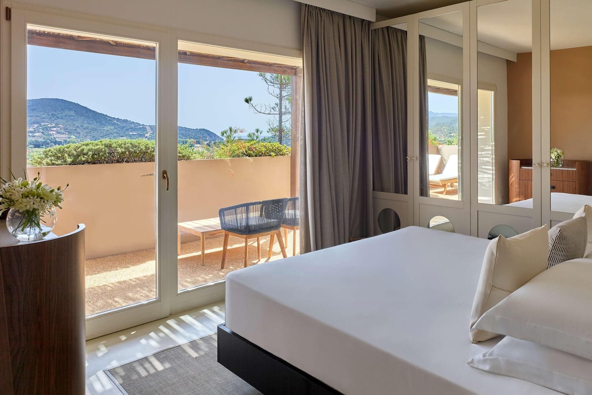 A hotel room with a large bed, a balcony with a view of the hills, a desk with a chair, and a mirror.