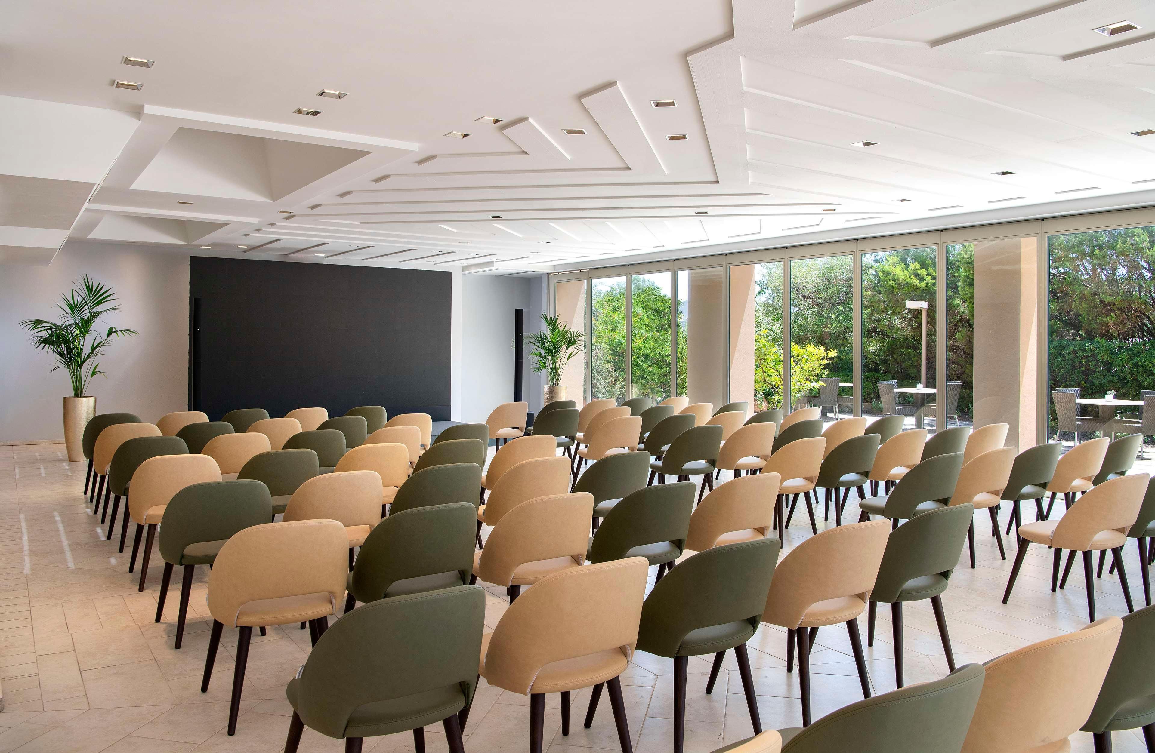 Une salle de conférence avec des rangées de chaises, un grand écran et de grandes fenêtres donnant sur un paysage verdoyant.