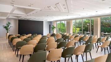 Une salle de conférence avec des rangées de chaises, un grand écran et de grandes fenêtres donnant sur un paysage verdoyant.