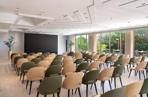 Un salón de conferencias con filas de sillas, una pantalla grande y amplias ventanas con vistas a zonas verdes.