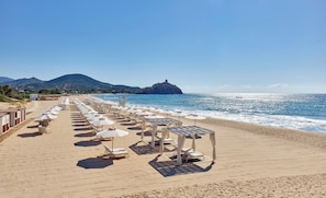 Una playa con filas de tumbonas y sombrillas, vista despejada al mar, y una montaña al fondo.