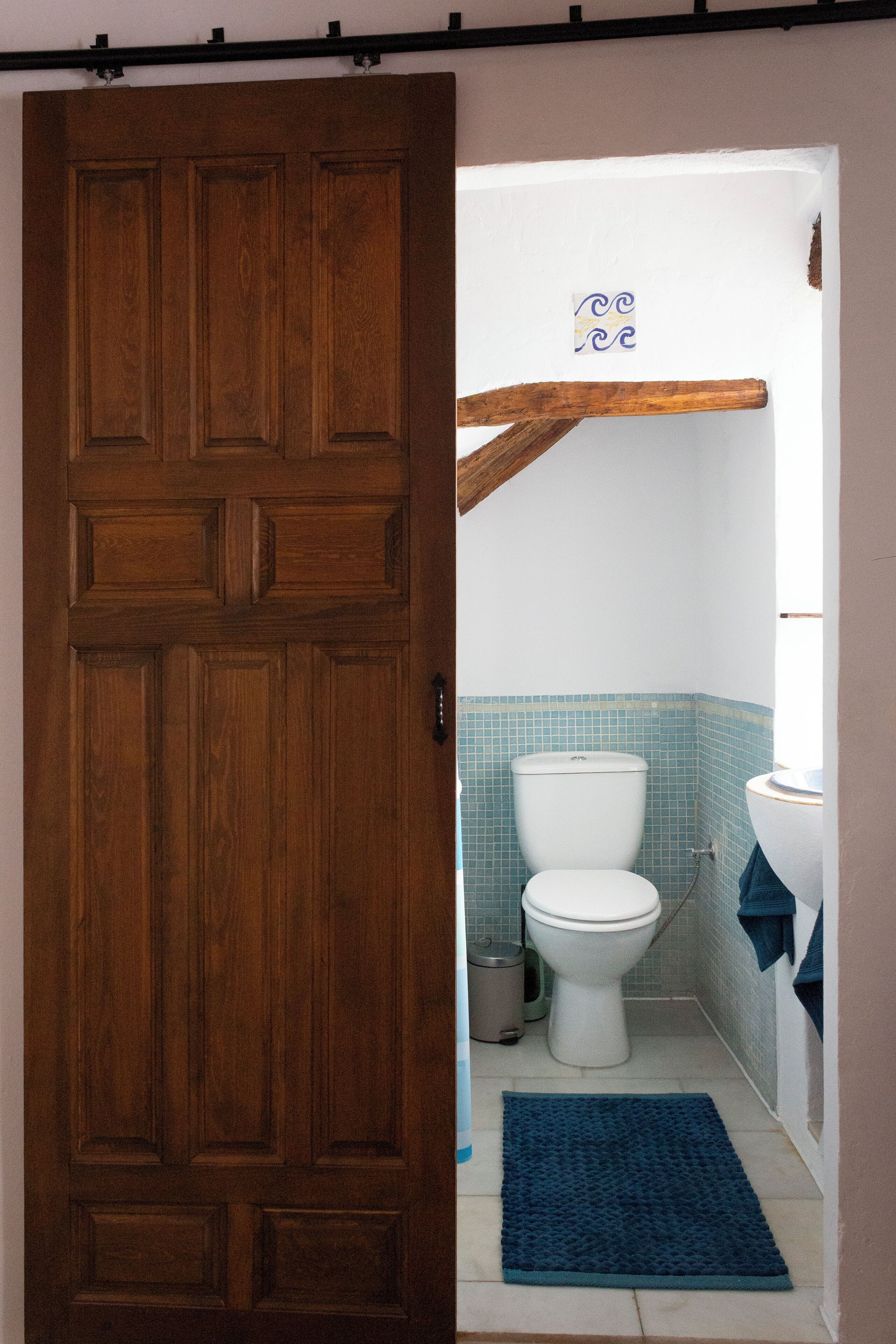 A wooden door leading to a bathroom with a toilet, sink, and blue tiles.
