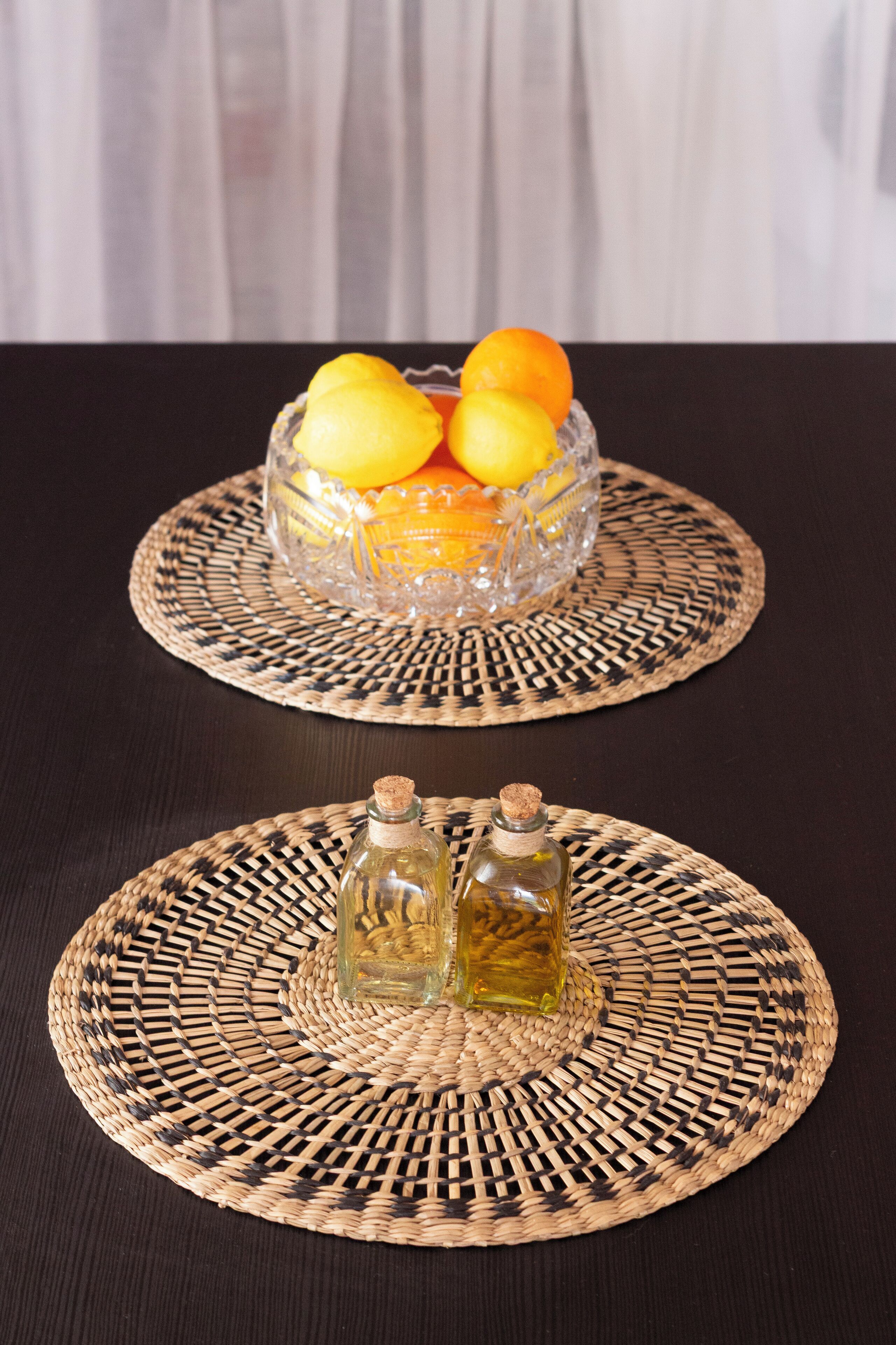 A table with a woven placemat and a glass bowl containing fruit.