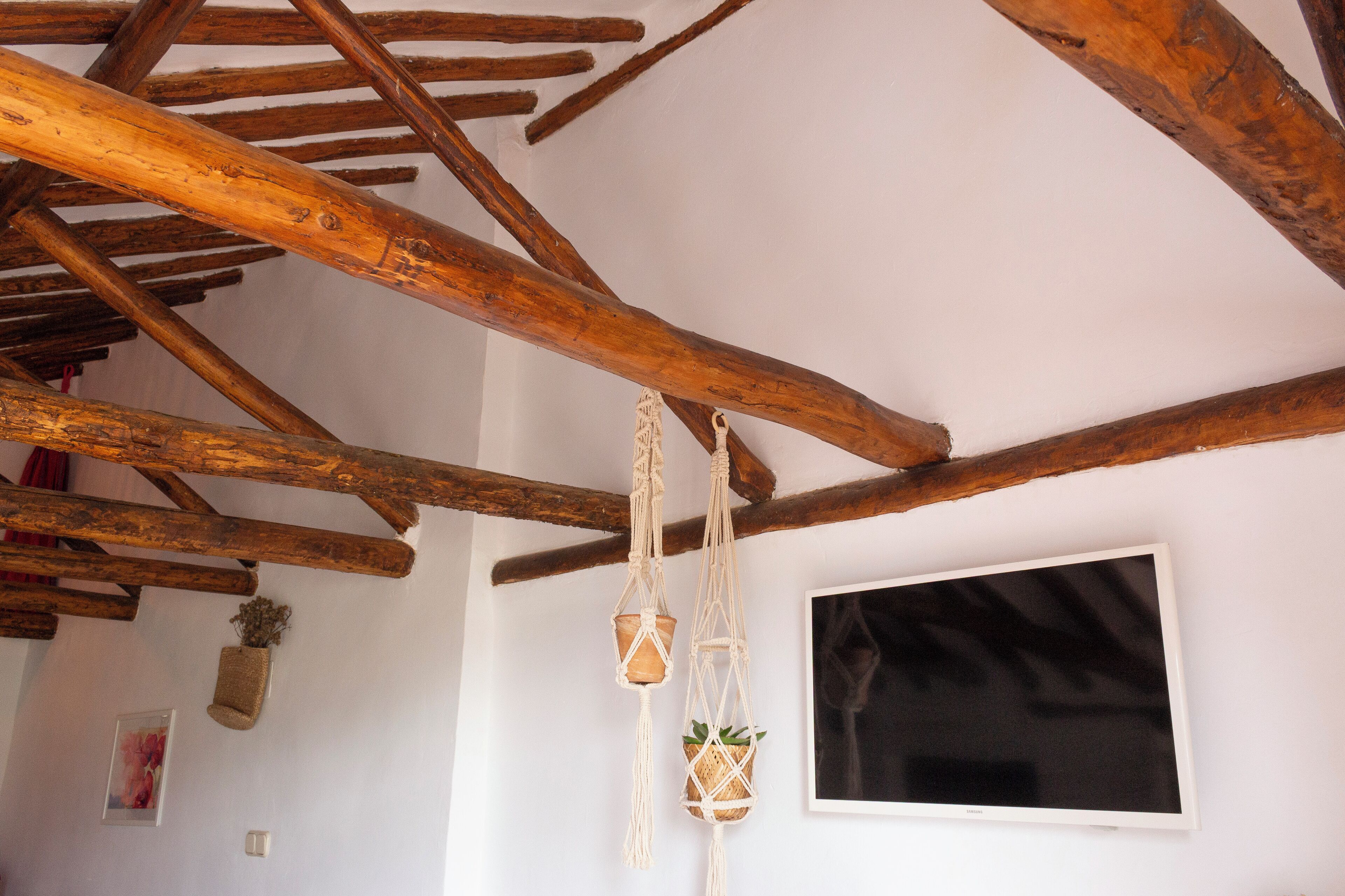 Un interior rústico con vigas de madera a la vista, una pared blanca, un cuadro enmarcado y tapices de macramé.