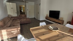 Un salon moderne avec une table à manger en bois, des chaises blanches, un canapé d’angle et un ventilateur de plafond.