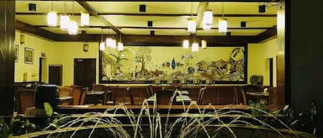 A restaurant with a long counter, hanging lights, and a large painting on the wall.