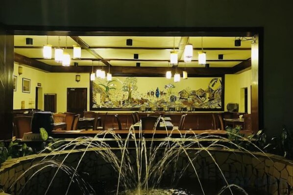 A restaurant with a long counter, hanging lights, and a large painting on the wall.