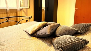 A bed with a patterned bedspread and several pillows, a black wardrobe, and a wooden door in a room with a window and a radiator. - Apartment in the center of Serravalle at 5min from the outlet Serravalle Mcarthurglen (Serravalle Scrivia)