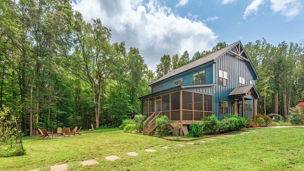 A two-story blue cabin with a deck and a covered porch, surrounded by a well-maintained lawn and trees.
