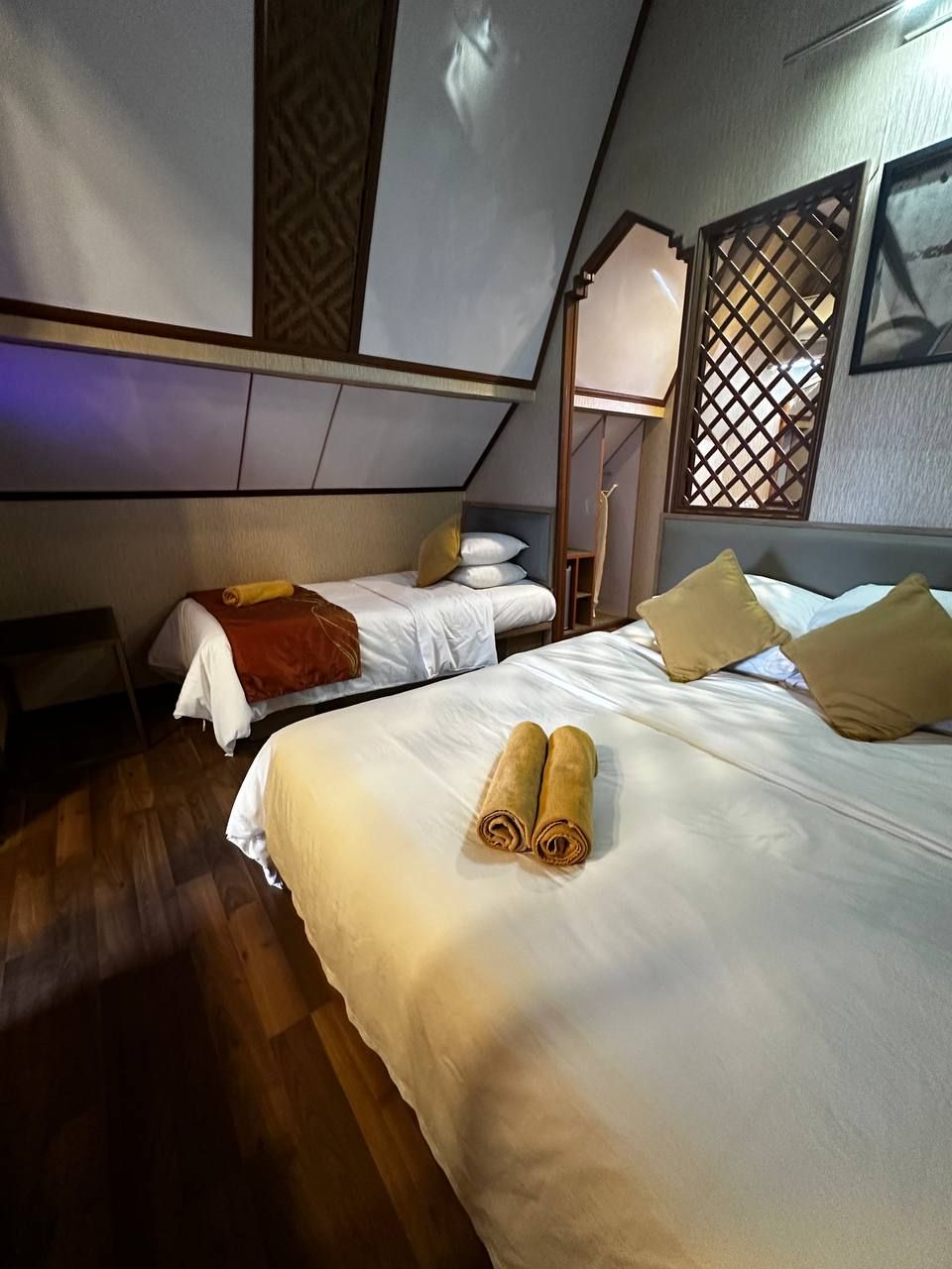 A hotel room with two beds, wooden flooring, and a window with a lattice design.