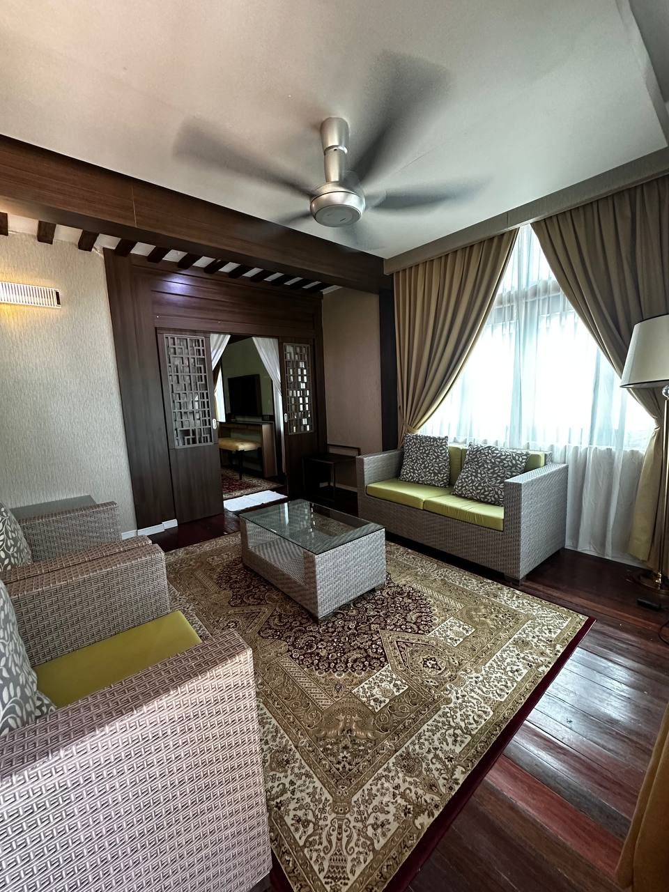 A modern living room with a glass coffee table, patterned sofas, a large rug, and a ceiling fan.
