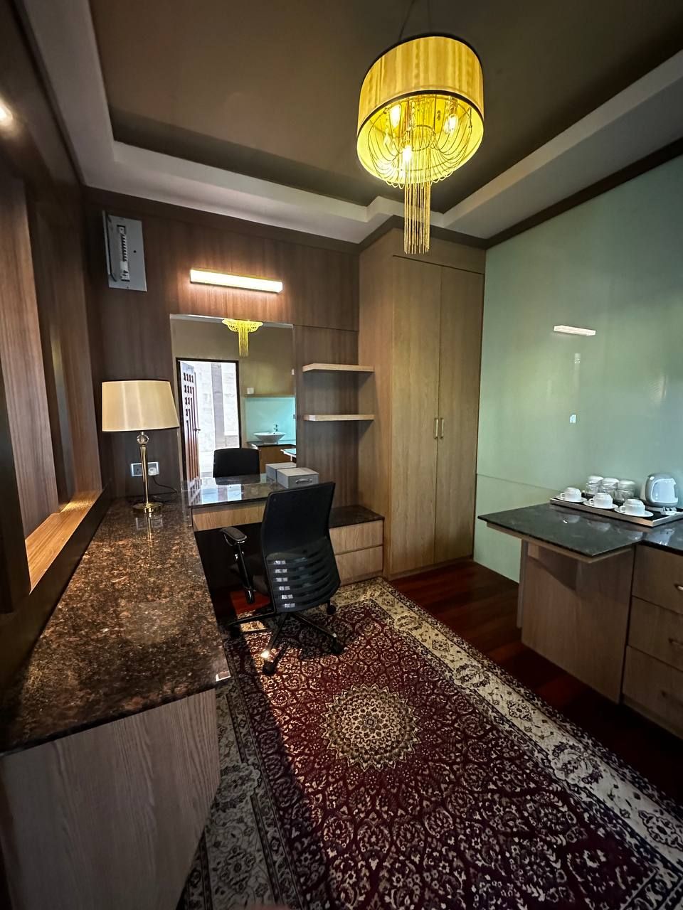 Una habitación de hotel moderna con encimera de mármol, una silla de oficina negra, alfombra estampada, armario de madera y candelabro.