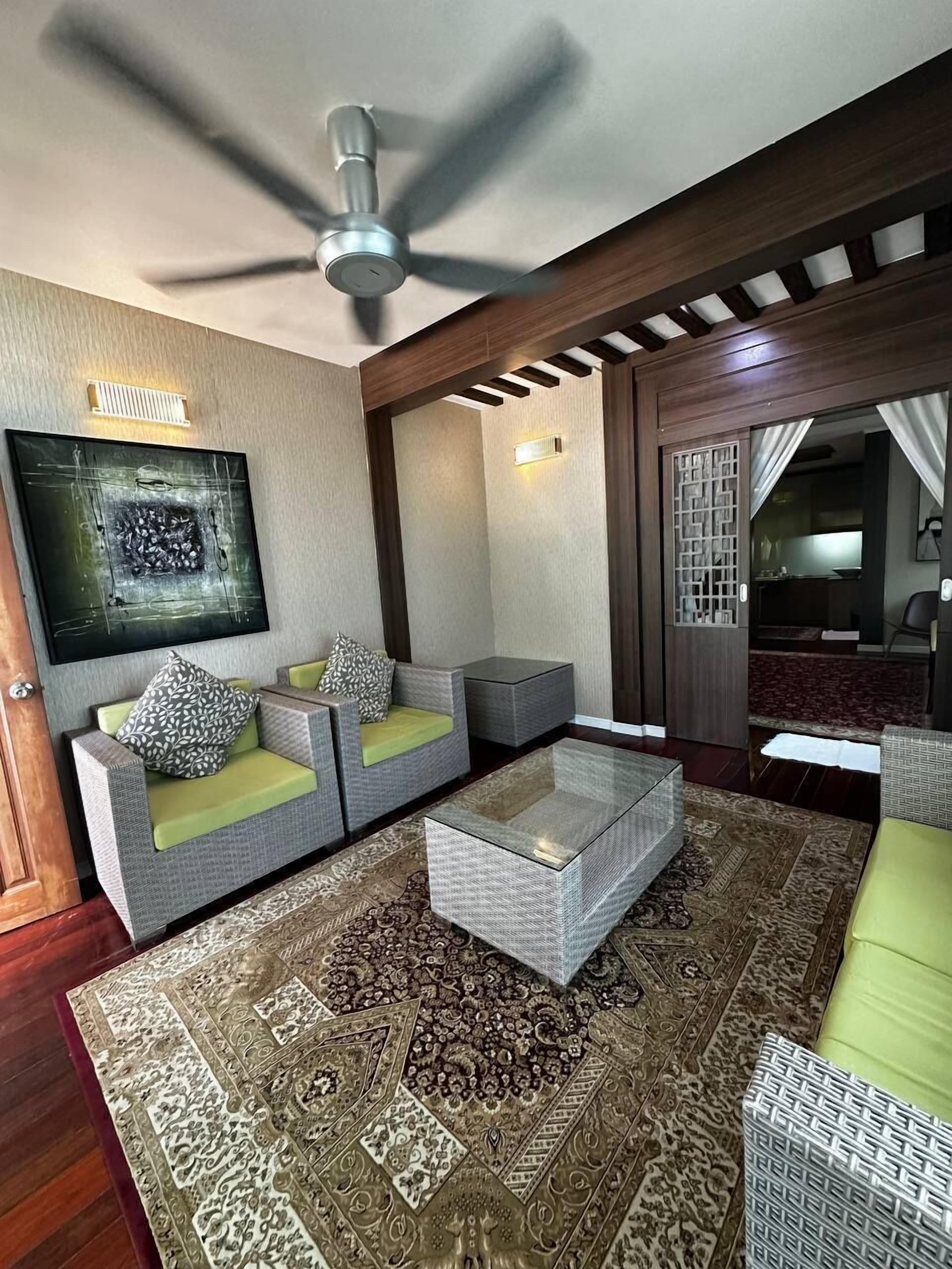 Una sala de estar moderna con ventilador de techo, dos sofás grises con cojines verdes, una mesa de centro de vidrio y un tapete estampado.