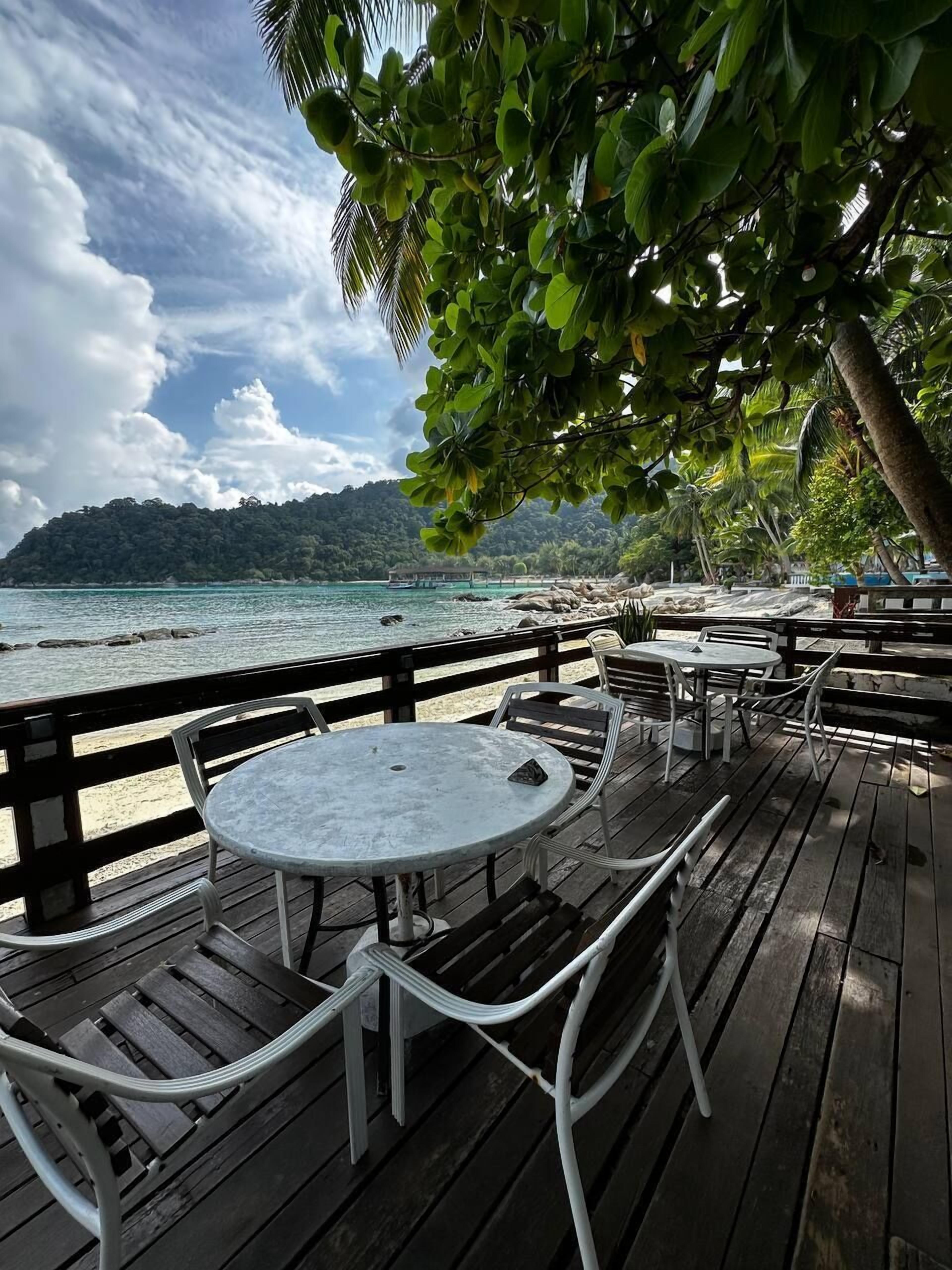 A wooden deck with tables and chairs overlooking a beach with clear water and a distant mountain.