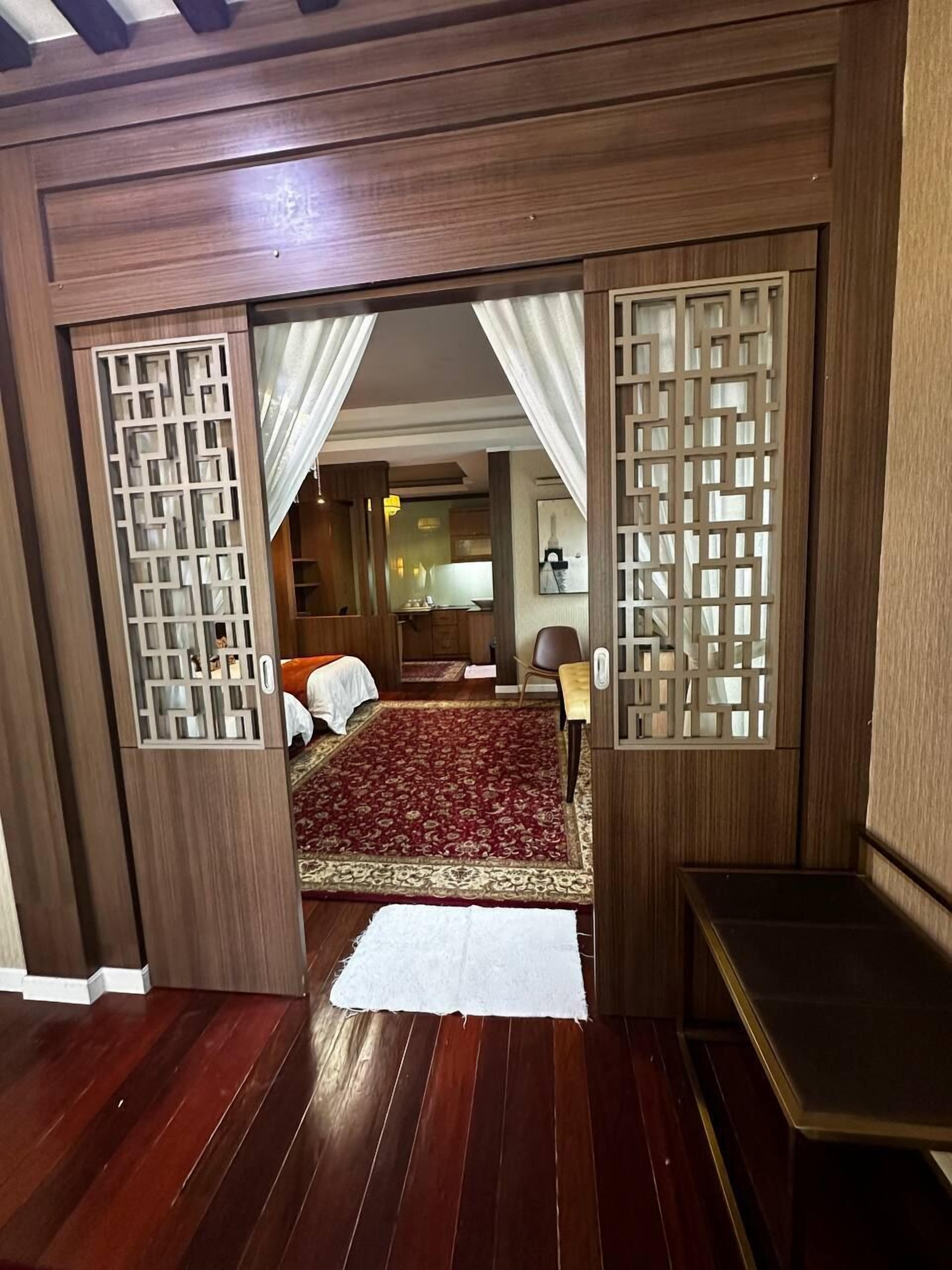 Una habitación con piso de madera, una cama con colcha estampada roja, un tapete blanco y un armario de madera.