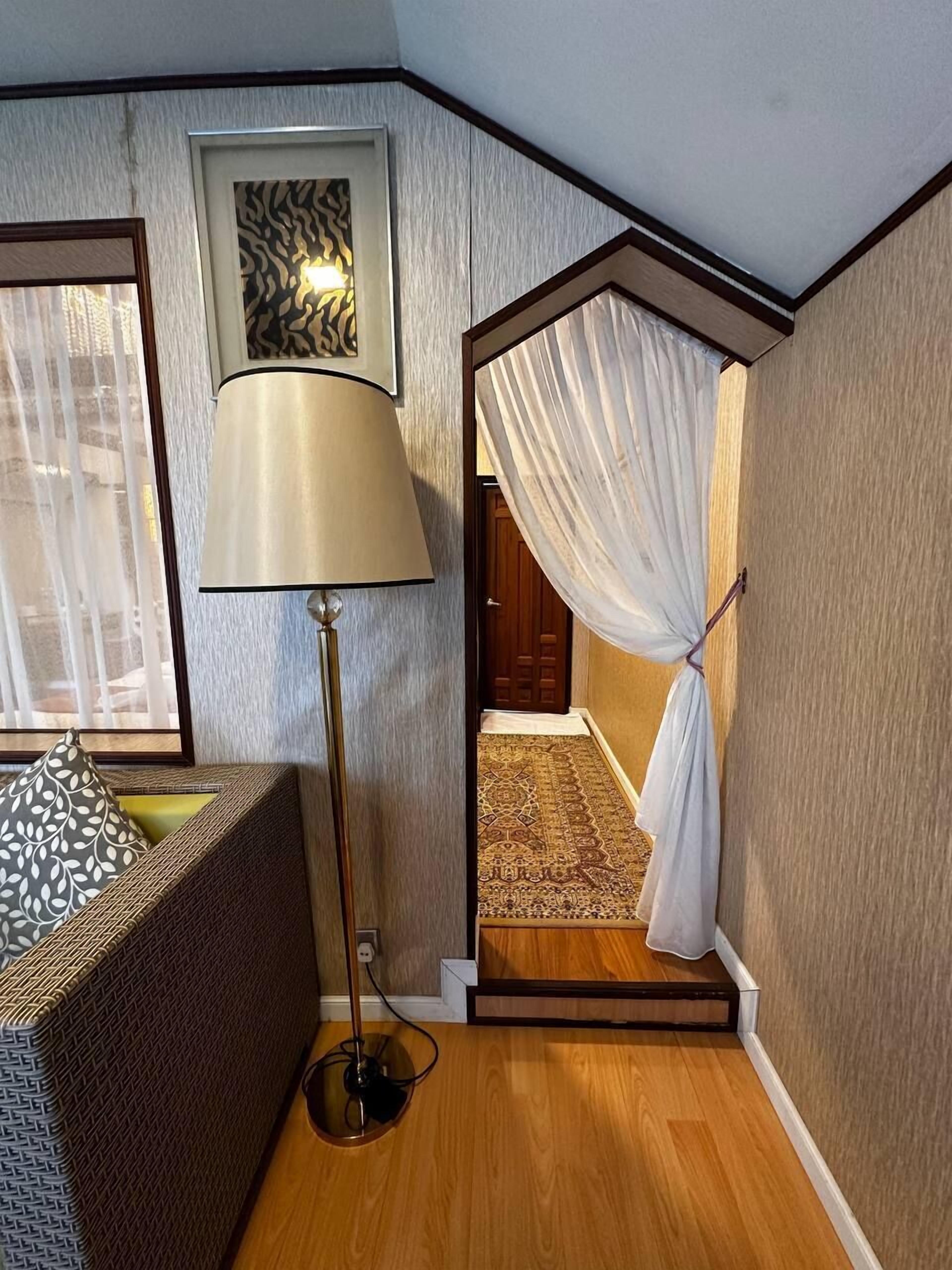 Une chambre d’angle avec un lampadaire, un tapis à motifs, un lit agrémenté d’un coussin décoratif et une porte donnant sur une autre pièce.