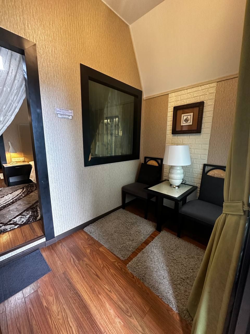 A compact room with a wooden floor, a small table with a lamp, a chair, and a framed picture on the wall.