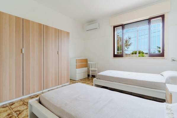 A bedroom with two beds, a wardrobe, a desk, and a window with a view of greenery.