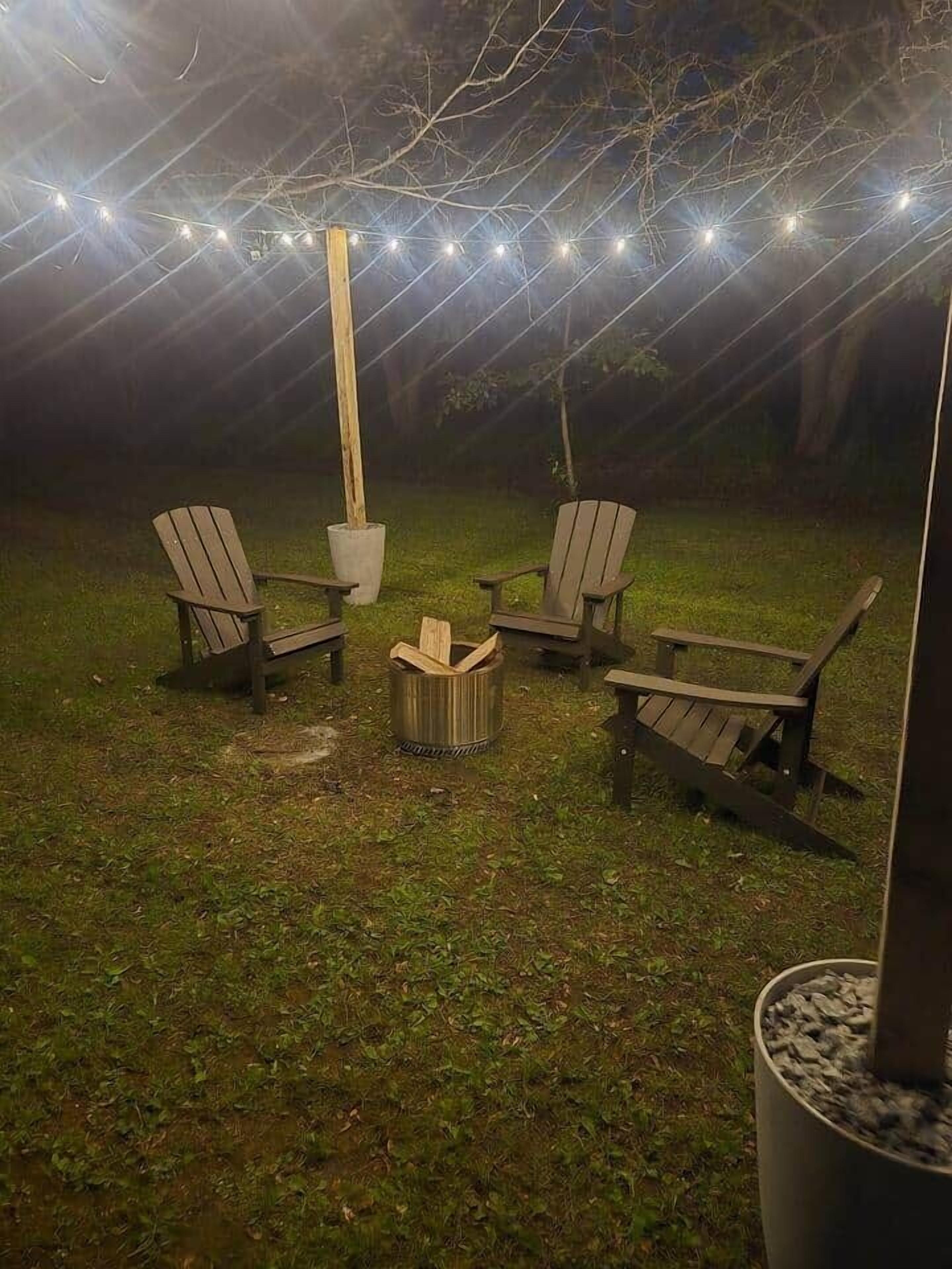 Un espace extérieur aménagé avec quatre fauteuils Adirondack, un foyer et des guirlandes lumineuses.