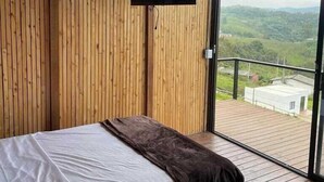A bedroom with a bed, a mounted TV, and a balcony with a view of greenery. - Samadhi Glamping (El Penol)