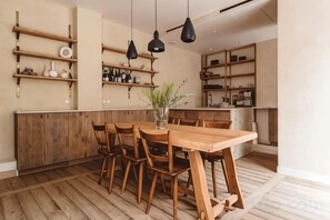 A dining area with a wooden table and chairs, hanging pendant lights, and wooden shelves with various items.