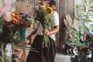 Una persona sostiene un ramo de flores en una habitación con diversas plantas y una mesa de madera.