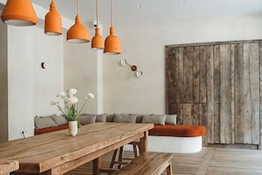 Un comedor con mesa de madera, bancos, lámparas colgantes naranjas y un florero blanco con flores.