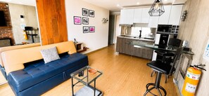 Uma sala de estar moderna com um sofá azul, uma mesa de centro de vidro e uma área de cozinha com armários brancos e bancada preta.