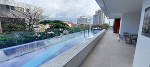 Ein Pool auf dem Dach mit Glasgeländer, Sitzgelegenheiten im Freien und Blick auf Stadtgebäude.