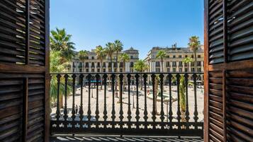 Ein Balkon mit schmiedeeisernem Geländer, der auf einen öffentlichen Platz mit Palmen und historischen Gebäuden blickt.