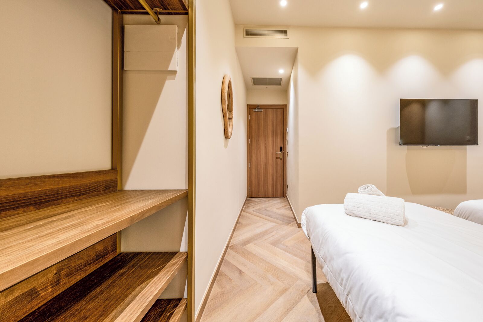 Une chambre d’hôtel avec un banc en bois, un lit recouvert d’une couverture blanche, un téléviseur fixé au mur et, au fond, une porte en bois.