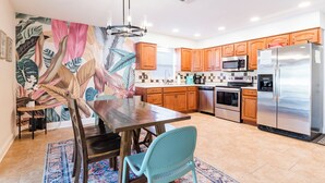 A modern kitchen with wooden cabinets, stainless steel appliances, a dining area with a table and chairs, and a colorful mural on the wall. - Heated Private Pool + Game Room | Family & Group Favorite | Beach Minutes Away (Largo)
