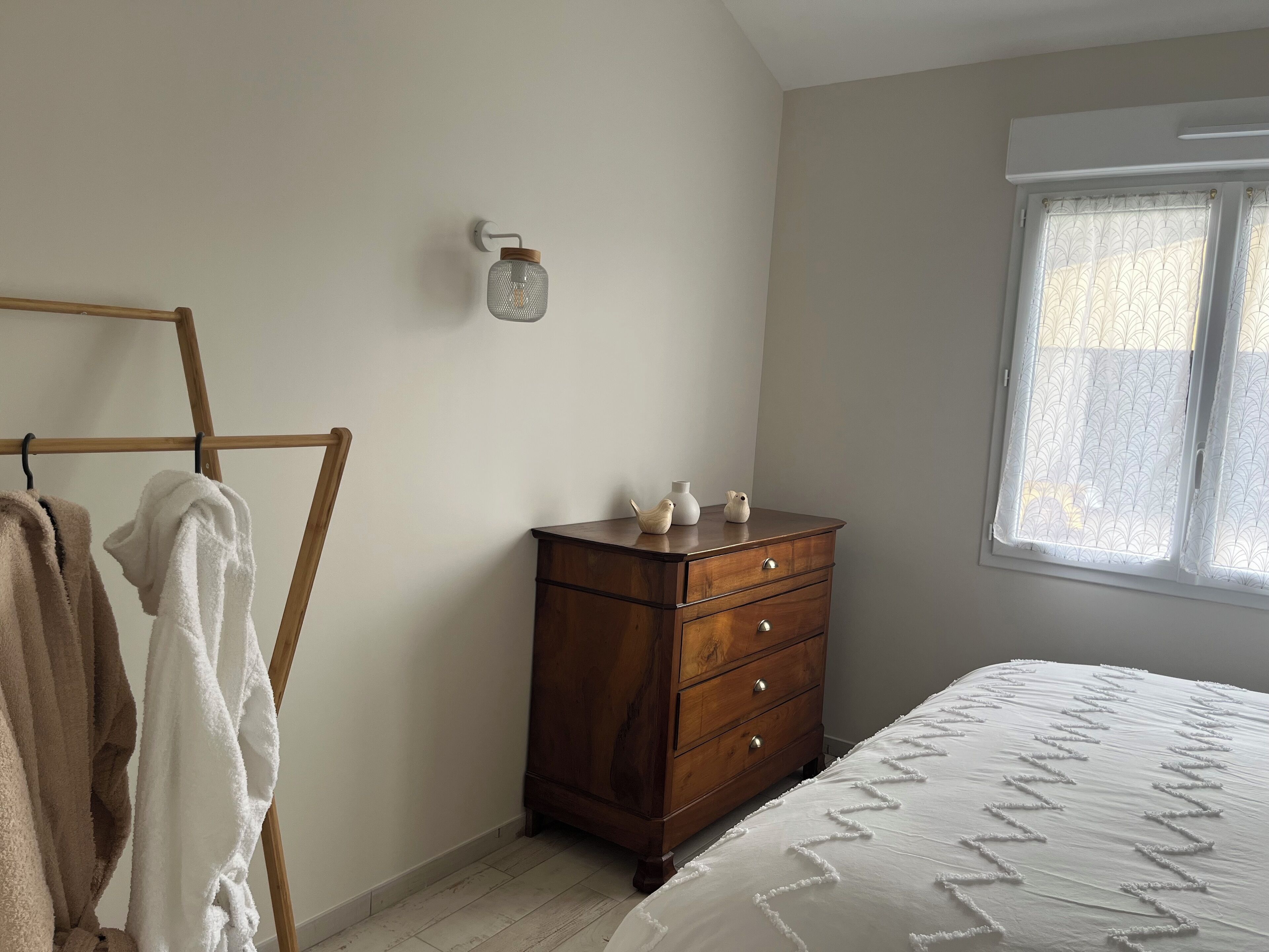 Une chambre avec une commode en bois, un lit recouvert d’une courtepointe blanche, une chaise en bois et un luminaire fixé au mur.