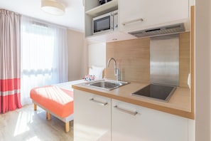 A modern kitchen with a microwave, sink, and stove, a bed with a red blanket, and a window with sheer curtains.